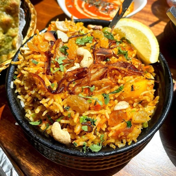 Close-up of Indian biryani with cashews, fried onions, and a lemon wedge in a black bowl.