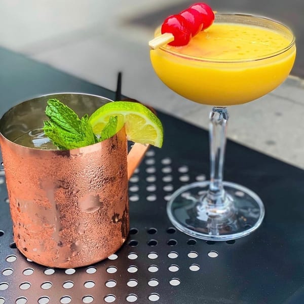 Two cocktails on a black table: a copper mug with mint and lime, and a yellow drink with cherries.